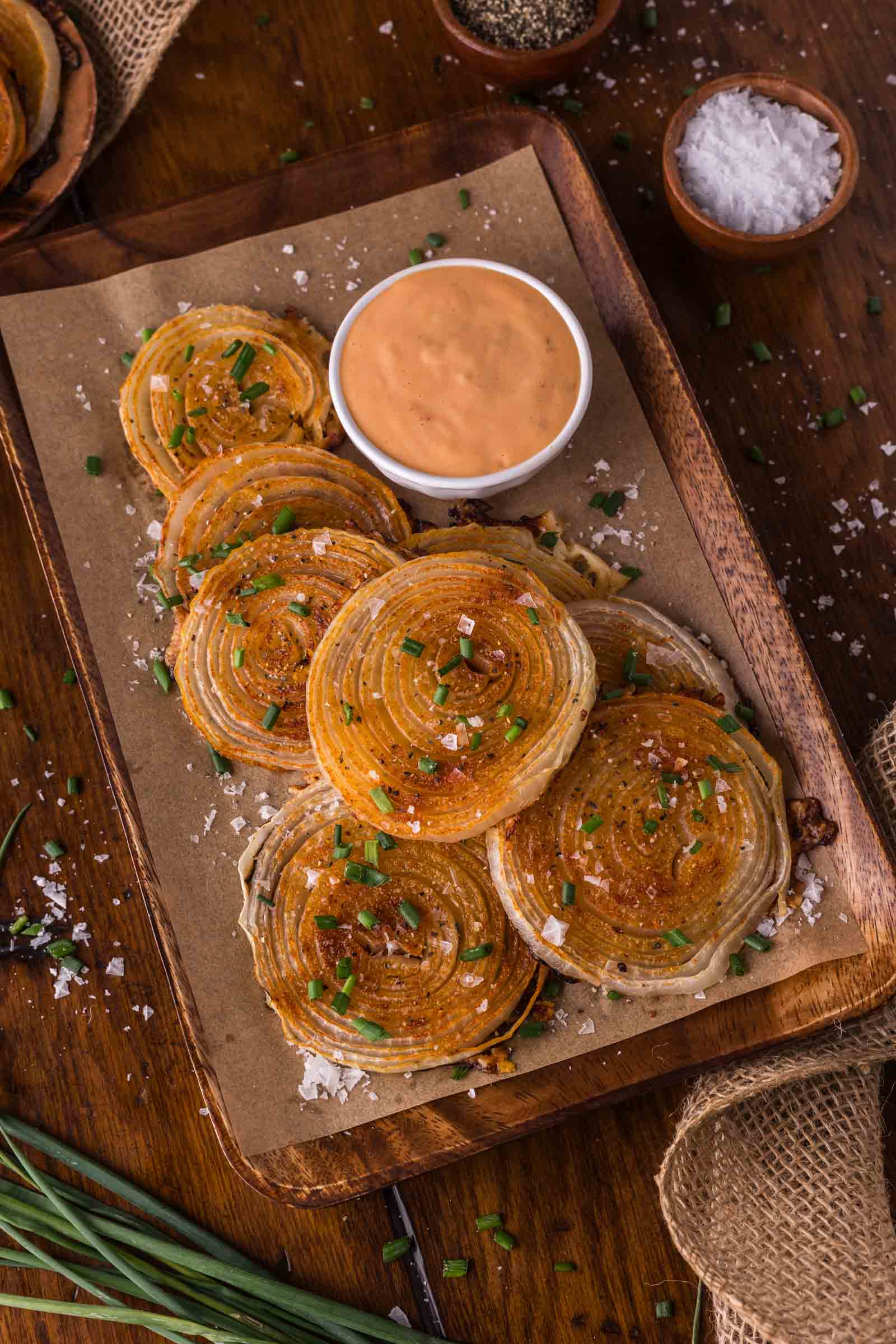A wooden tray with crispy roasted onion chips topped with chopped chives and coarse salt, served with a bowl of creamy dipping sauce.