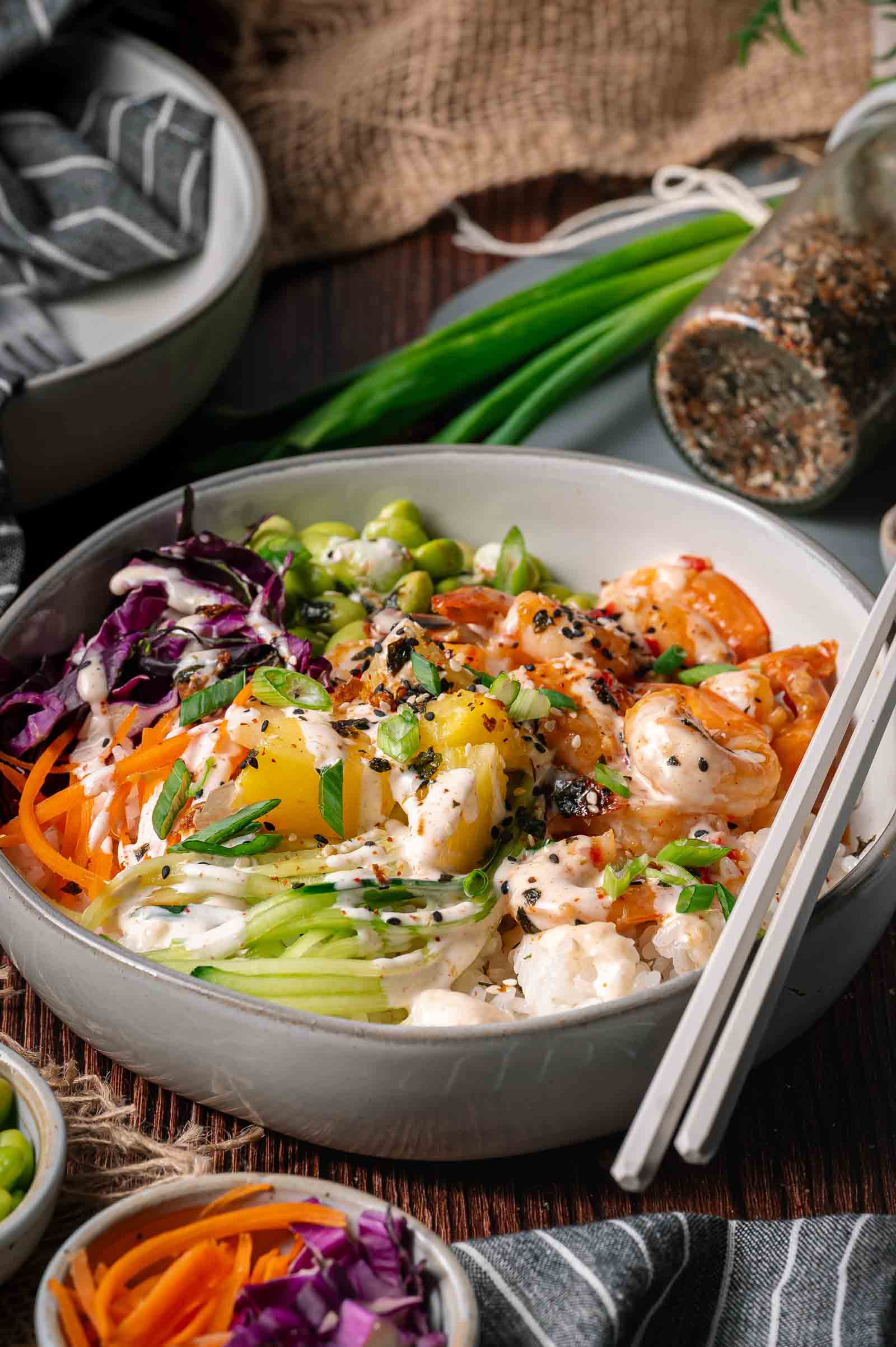 A poke bowl of rice topped with shrimp, pineapple, cucumber, carrot, edamame, red cabbage, and sesame seeds, served with chopsticks and a creamy dressing.