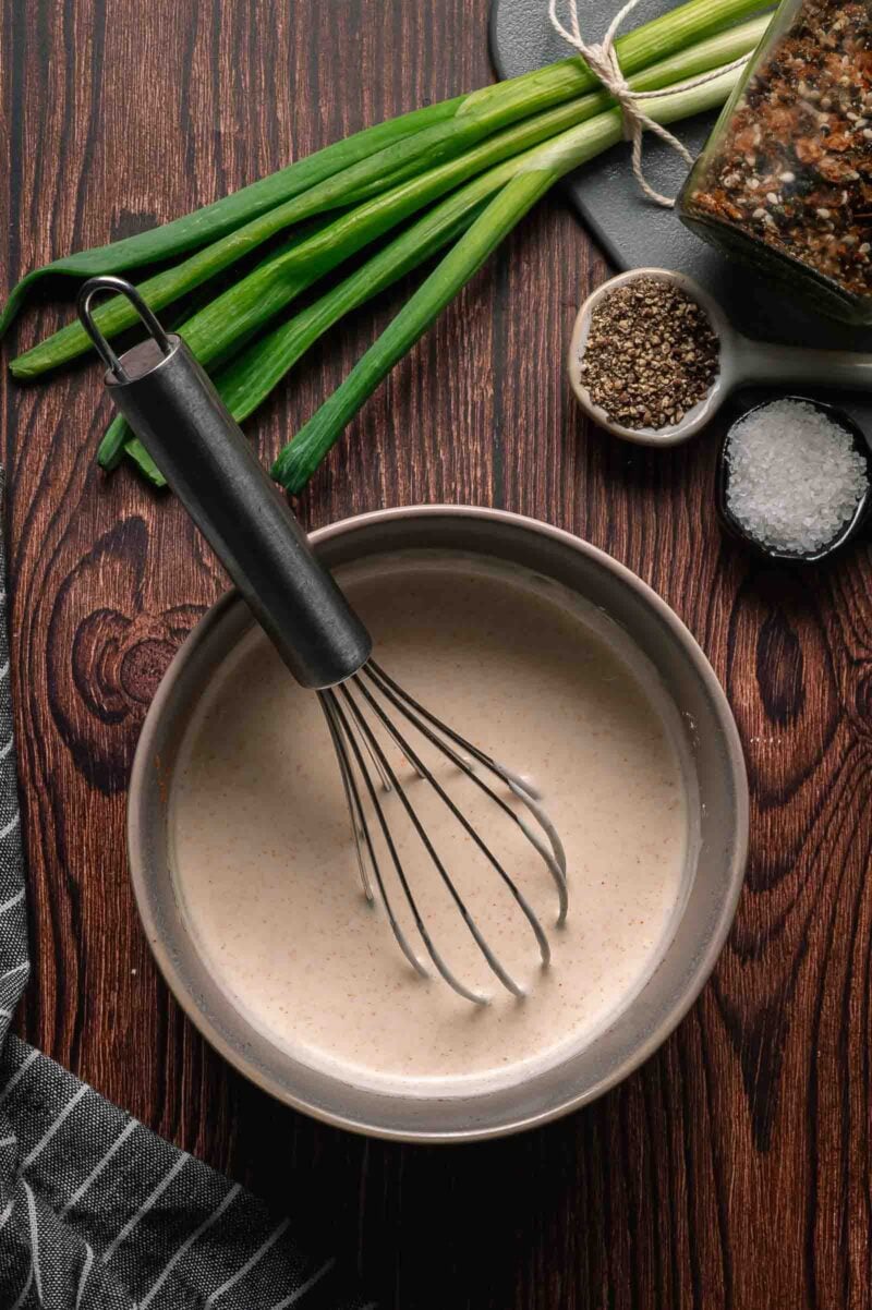 A bowl of creamy Japanese white sauce with a whisk, surrounded by green onions, a jar of spices, black pepper, and coarse salt on a wooden surface.
