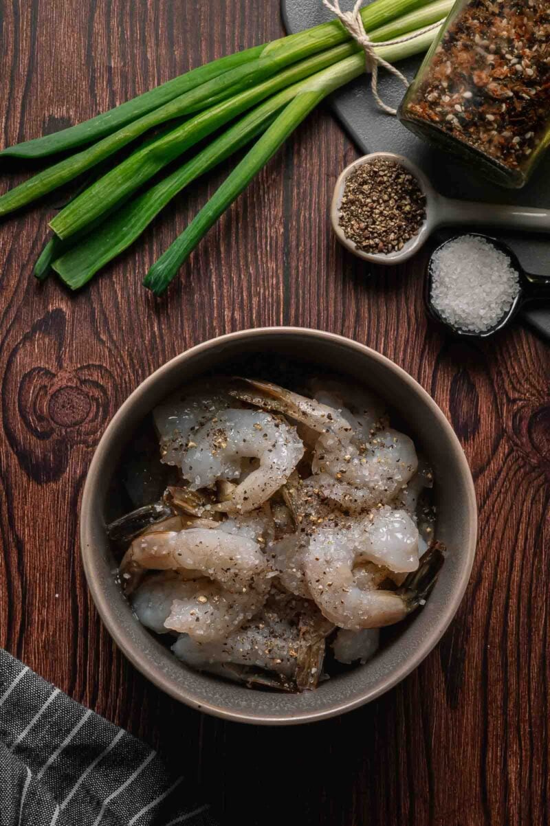 A bowl of raw shrimp seasoned with salt and pepper sits on a wooden surface, next to green onions, a spice jar, a dish of salt, and black pepper.