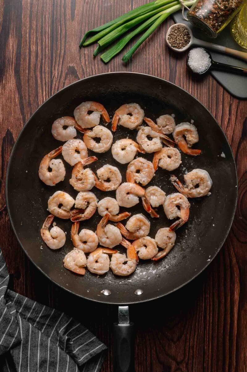 Shrimp cooking in a black skillet on a wooden surface, surrounded by green onions, salt, pepper, and seasonings.
