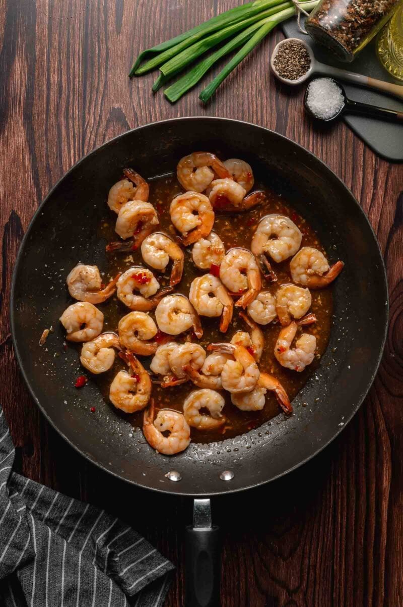 Shrimp cooked in a skillet with chili sauce, photographed from above on a wooden table next to green onions, black pepper, salt, and a striped kitchen towel.