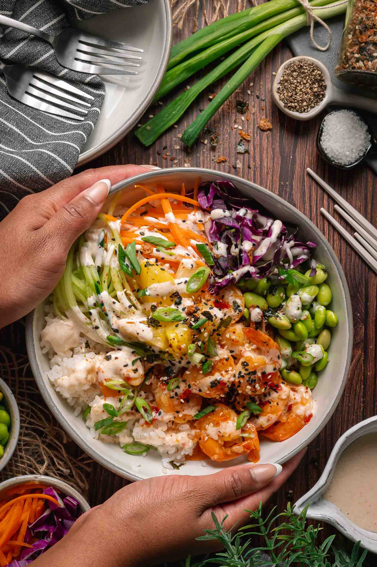 A person holds a poke bowl filled with rice, shrimp, shredded carrots, purple cabbage, edamame, cucumber, pineapple, and a drizzle of sauce, surrounded by fresh ingredients and utensils.
