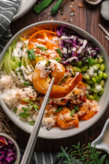 A close-up of chopsticks holding a cooked shrimp above a bowl filled with rice, shredded vegetables, edamame, and sauce.