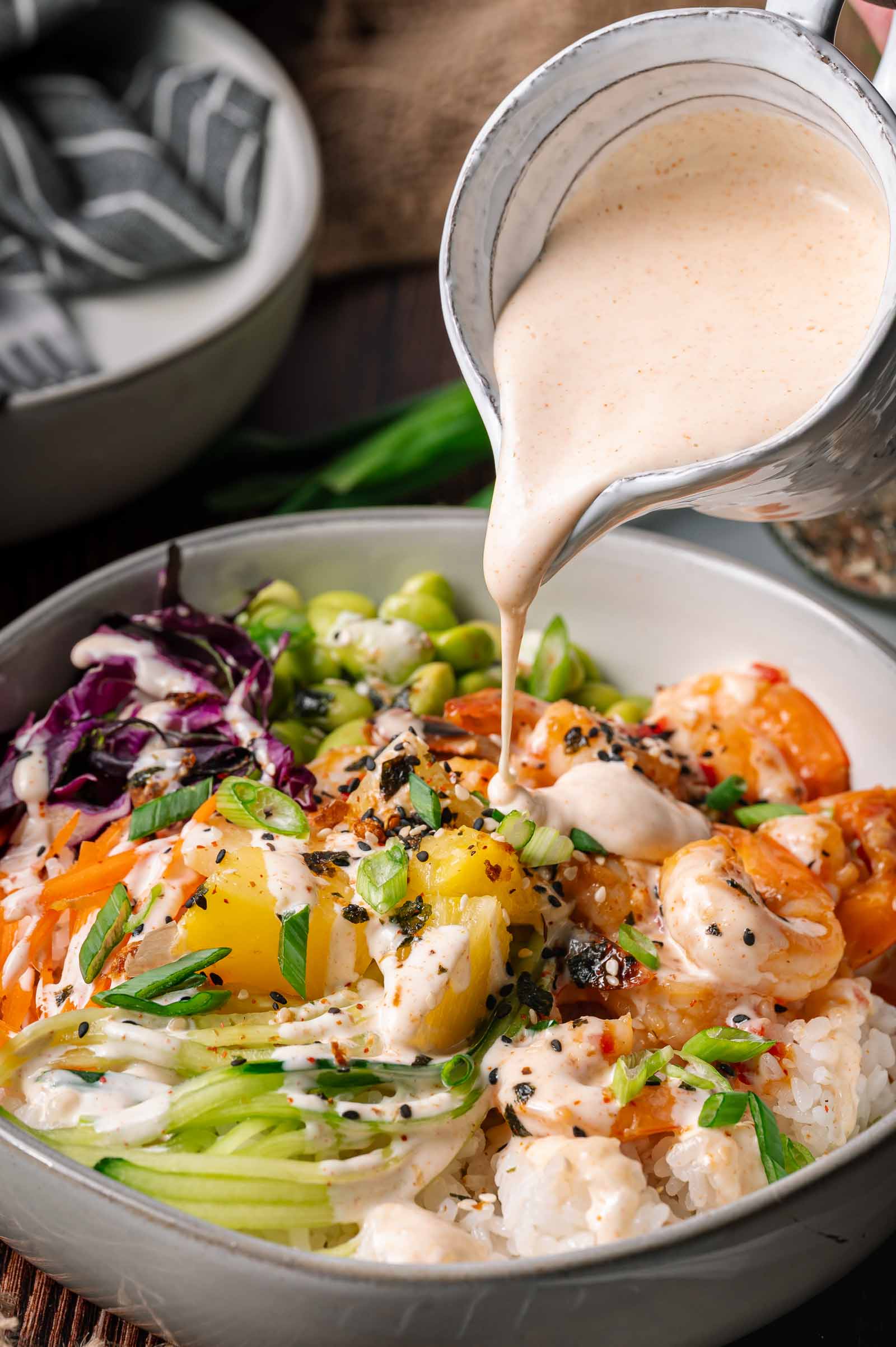 A bowl of rice topped with sweet chili shrimp, edamame, cabbage, pineapple, cucumber, and Japanese white sauce being poured over the dish.