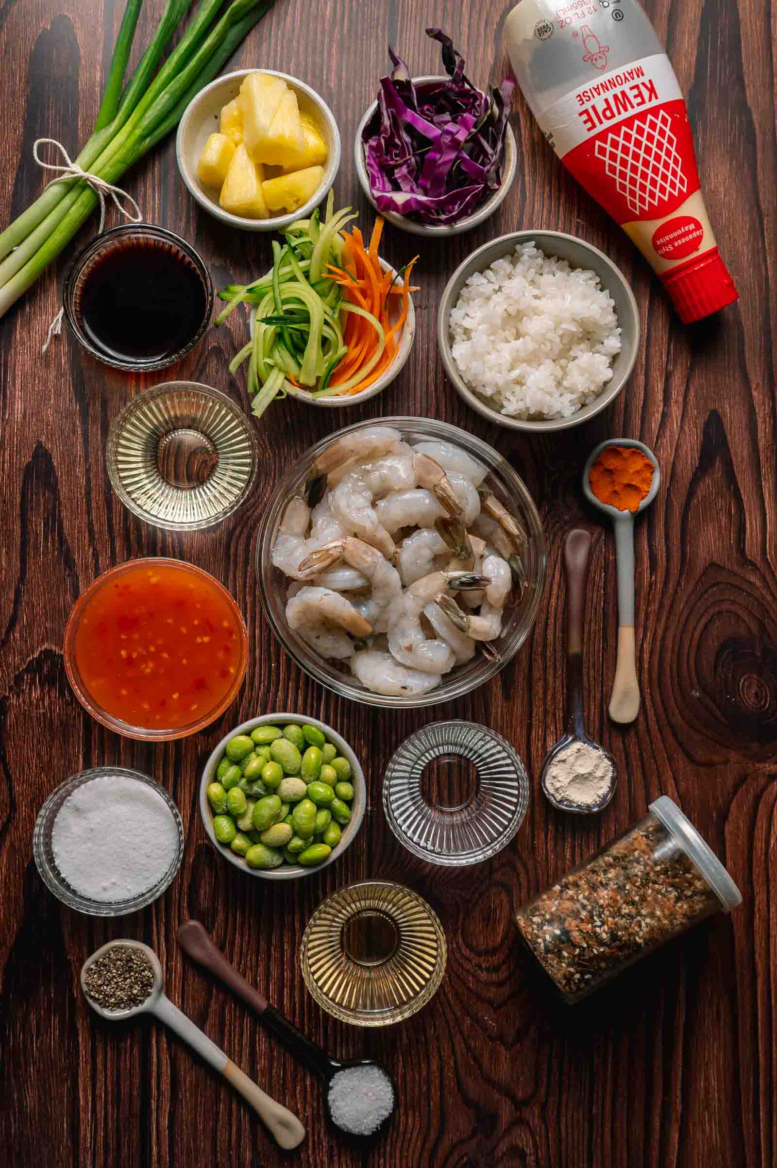 Assorted ingredients for a poke shrimp rice bowl, including raw shrimp, rice, vegetables, sauces, seasoning, pineapple, edamame, and Kewpie mayo, arranged on a wooden surface.