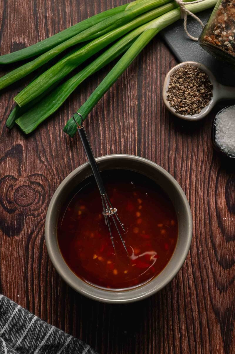 A bowl of reddish-brown chili sauce with a whisk, next to green onions, ground pepper, and coarse salt on a wooden surface.