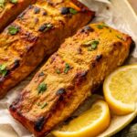 Three pieces of baked salmon fillet garnished with chopped herbs, served on a plate with lemon wedges and fresh greens.
