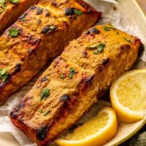 Three pieces of baked salmon fillet garnished with chopped herbs, served on a plate with lemon wedges and fresh greens.