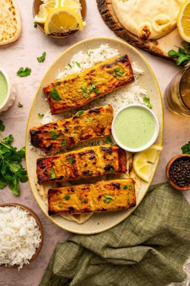 Four tandoori fish fillets on white rice, garnished with herbs, served with green sauce, lemon wedges, naan bread, and bowls of rice and pepper.