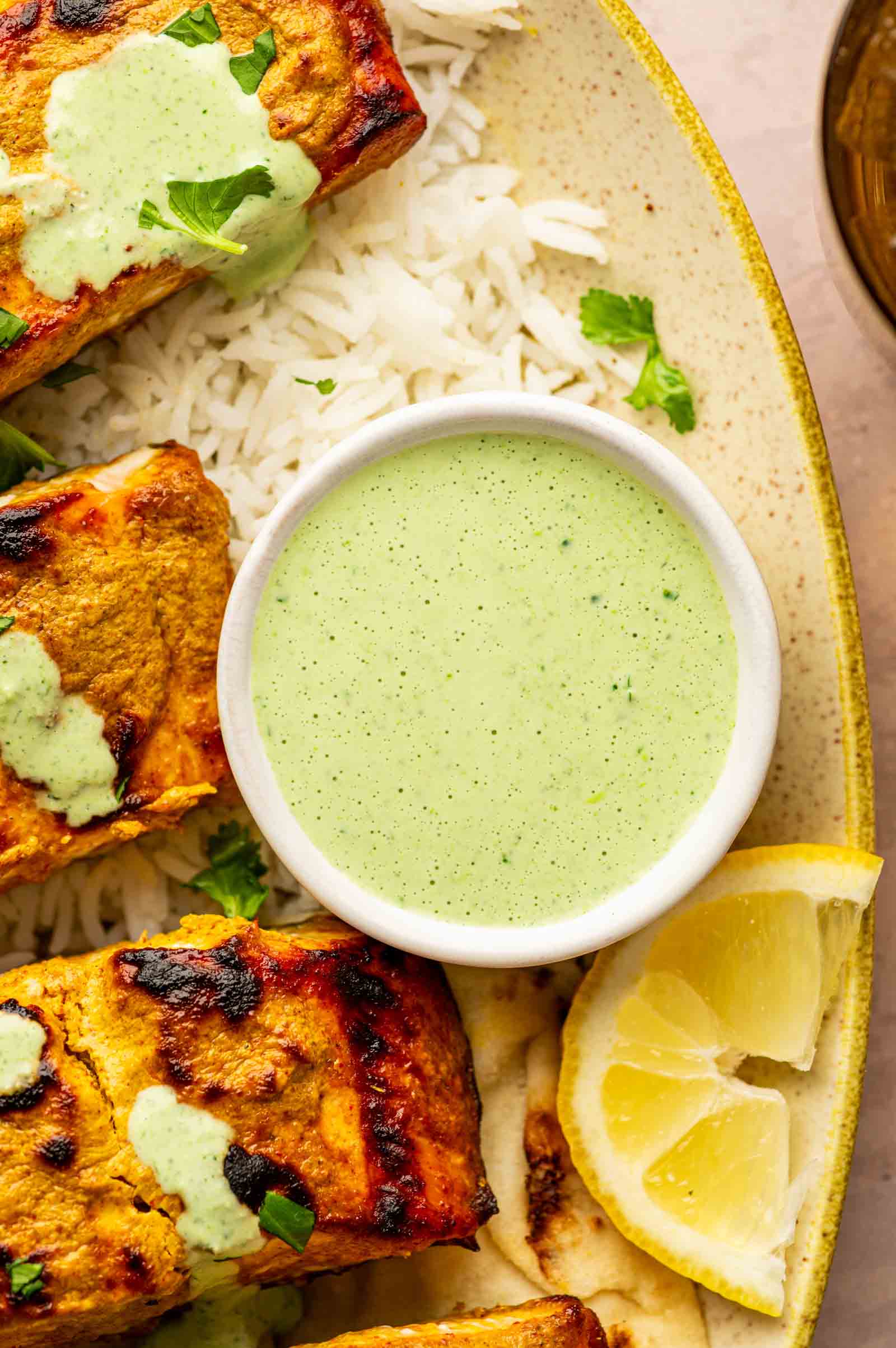 A bowl of green sauce sits on a plate with grilled marinated paneer, basmati rice, naan, and lemon wedges.