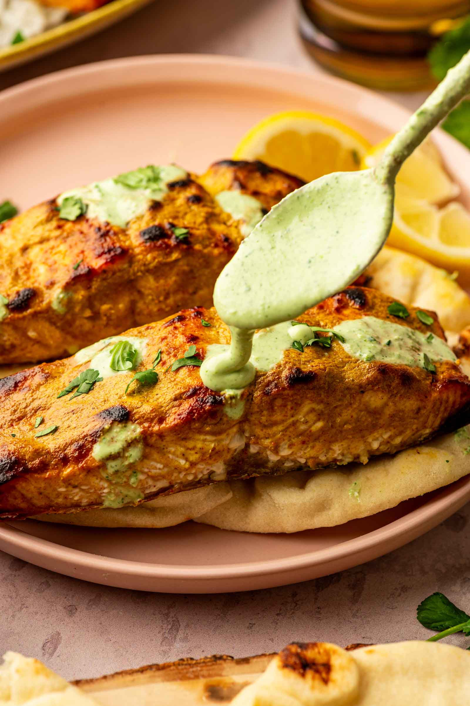 A spoon drizzles green sauce over two pieces of grilled salmon garnished with herbs, served on flatbread with lemon slices on a pink plate.