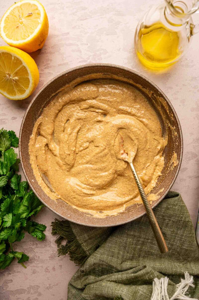 A bowl of creamy, light brown tandoori sauce with a spoon, surrounded by lemon halves, a bottle of oil, cilantro, and a green cloth.