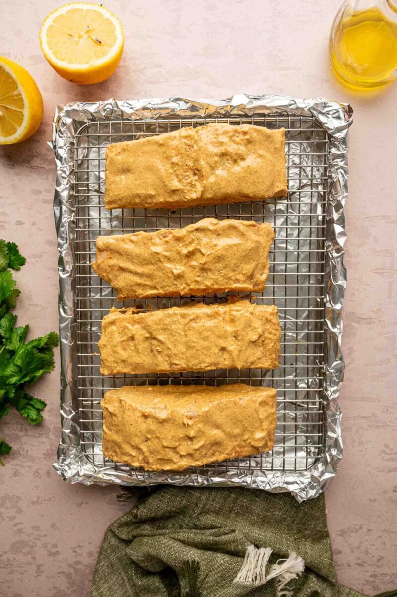 Four salmon fillets coated with a tandoori marinade mixture are placed on a wire rack over a foil-lined baking sheet, with halves of lemon, parsley, and a glass of oil nearby.