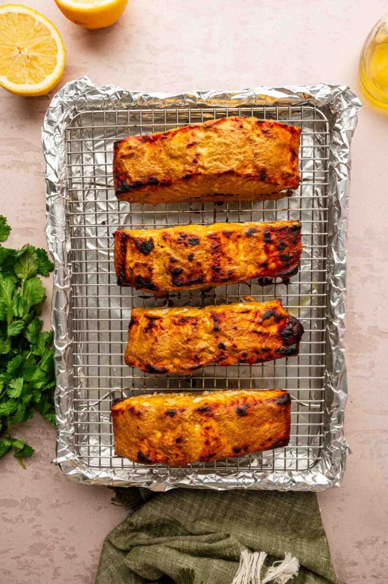 Four cooked salmon fillets on a wire rack over a foil-lined baking sheet, with lemon halves and fresh herbs nearby.