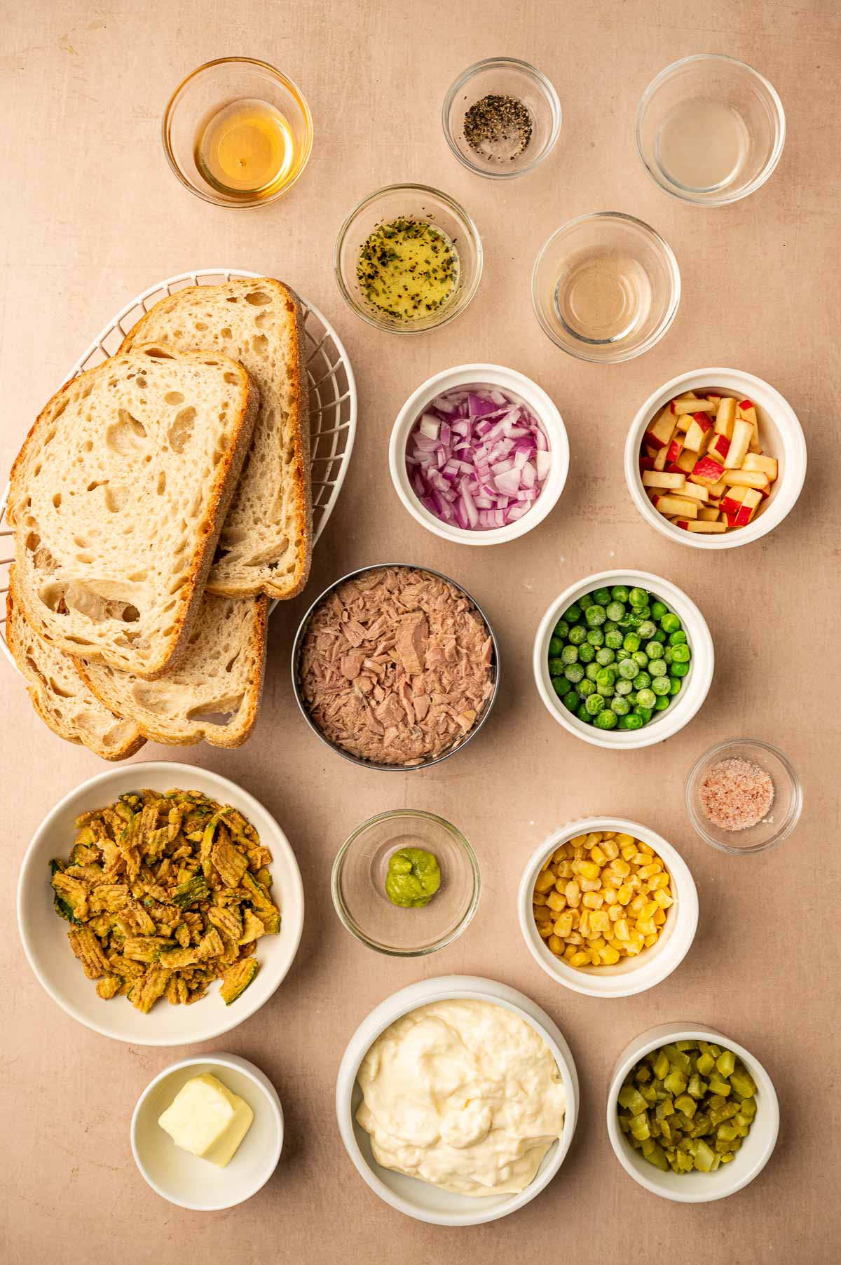Top-down view of various sandwich ingredients, including sliced bread, tuna, peas, onions, apples, corn, pickles, mayonnaise, seasonings, and small bowls of condiments on a beige surface.