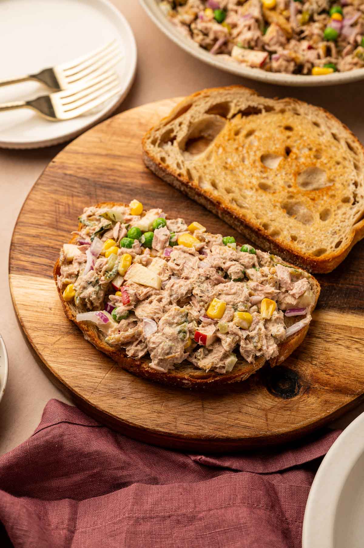 An open-faced tuna salad sandwich with corn, diced apples, and a slice of toasted bread on a wooden board, with plates and forks nearby.