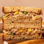 A close-up of a sandwich cut in half, filled with a mixture of tuna, corn, peas, and chopped vegetables on brown bread, resting on a wooden surface.