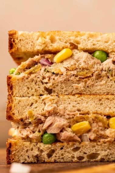 Close-up of two stacked tuna salad sandwiches with visible corn, peas, and red onion on rustic bread.