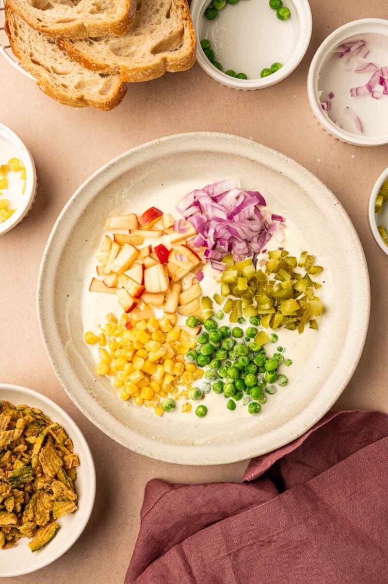 A bowl containing separate piles of chopped apples, red onion, pickles, corn, and peas on a creamy mayo base, surrounded by small bowls and sliced bread.