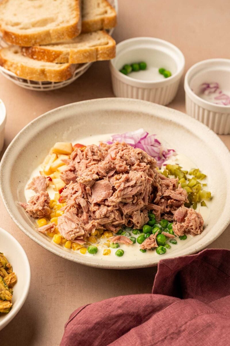 A bowl with tuna, chopped red onions, peas, sweetcorn, and diced jalapeños, surrounded by small bowls of ingredients and a plate of sliced bread in the background.