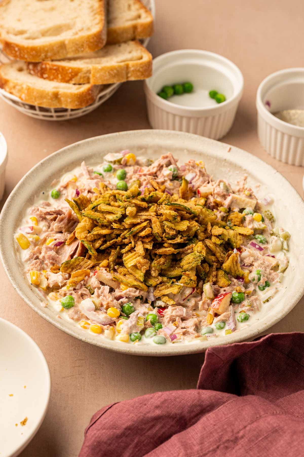A plate of mixed tuna salad topped with crispy potato chips, surrounded by slices of bread and small bowls on a beige surface.