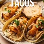 Three Baja fish tacos with battered fish, slaw, and sauce are served on tortillas with a side of dipping sauce, garnished with cilantro.
