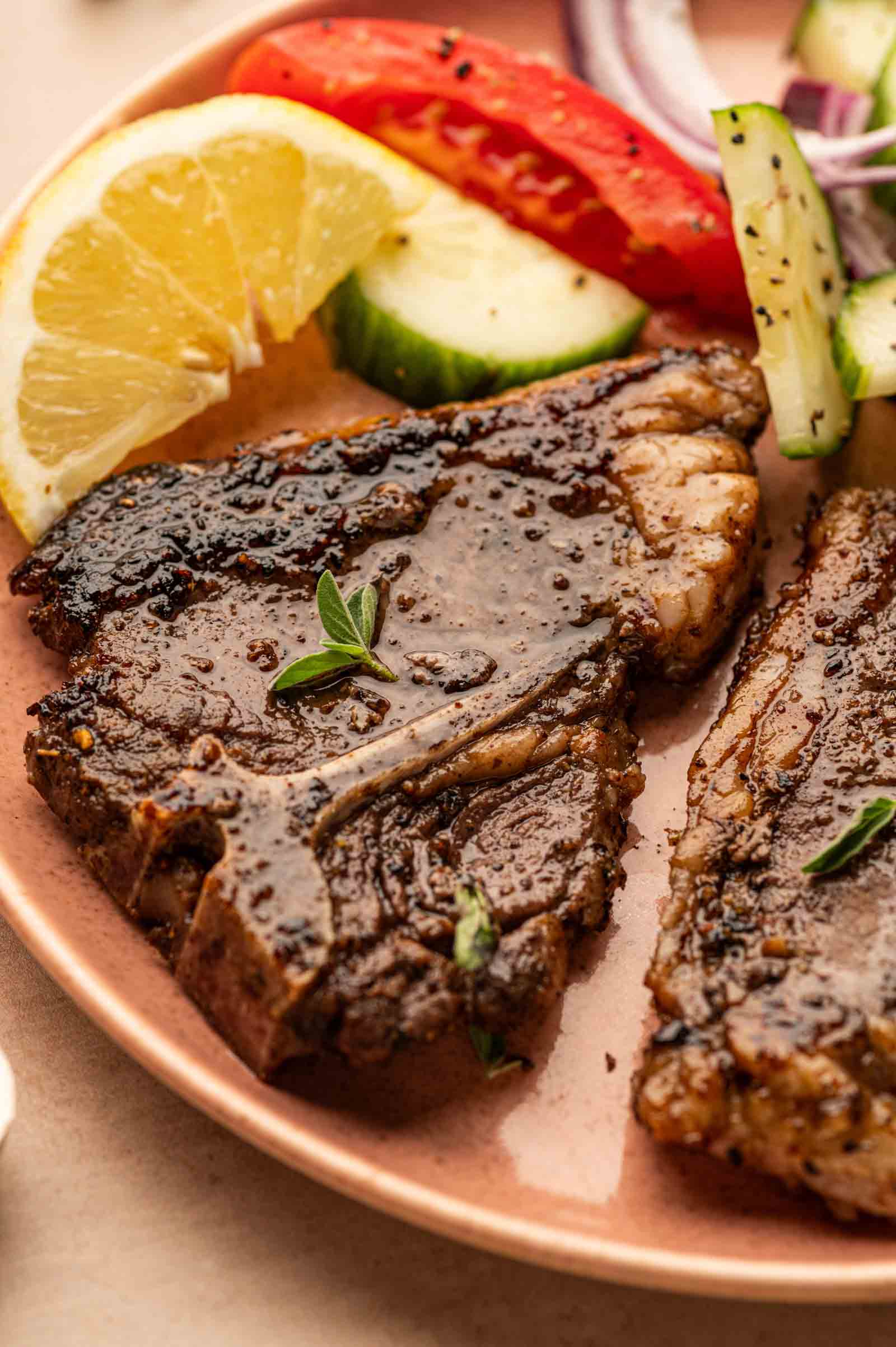 A Greek lamb chop served with lemon slices, cucumber, tomato, and red onion on a plate.