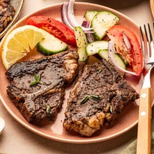 A plate with two cooked lamb chops, a lemon wedge, and a salad of tomato, cucumber, and red onion, with a fork and a green napkin beside the plate.