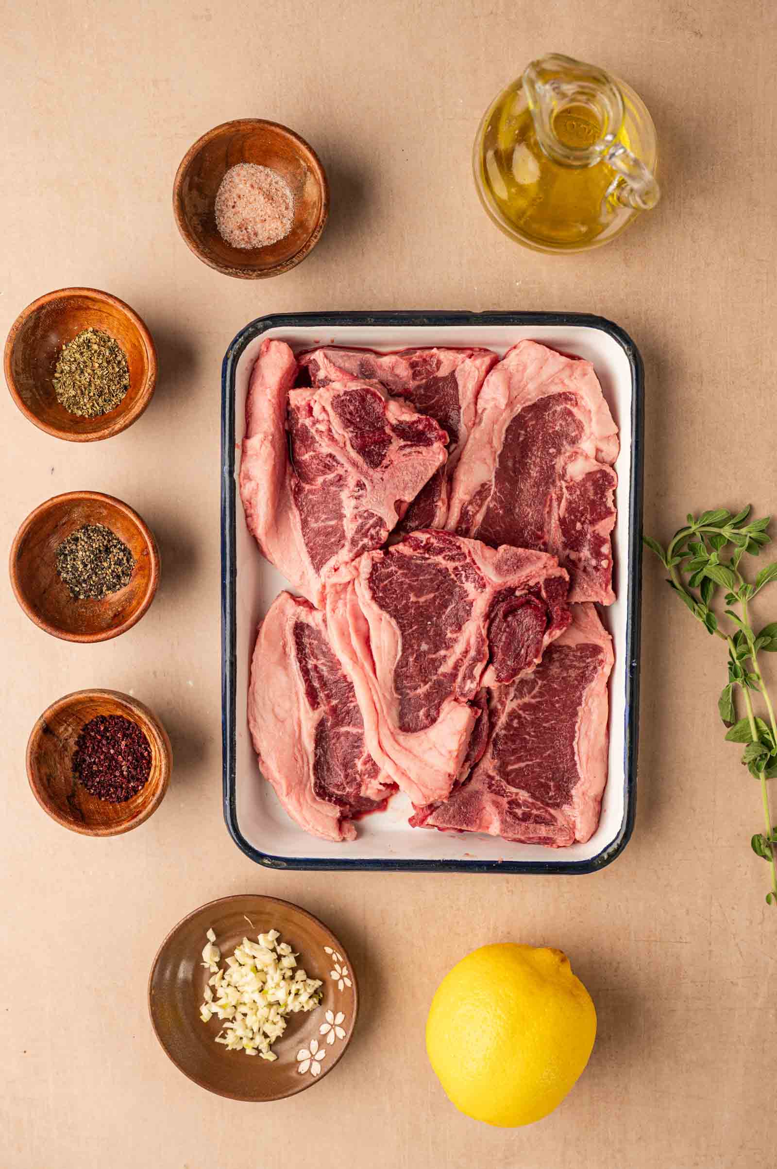 Raw lamb chops in a tray surrounded by bowls of spices, minced garlic, a lemon, fresh herbs, and a glass container of olive oil on a beige surface.