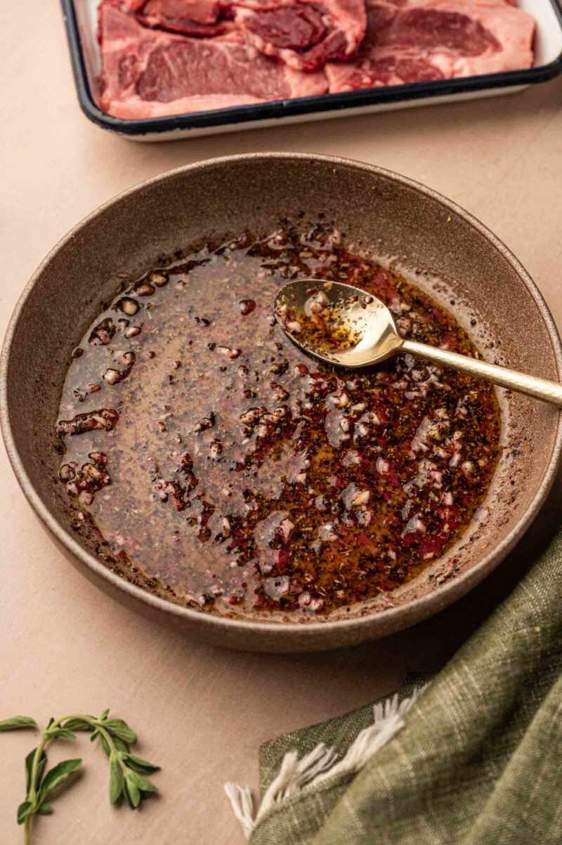A bowl of seasoned Greek lamb marinade with a spoon, sitting on a table next to a tray of raw meat and a green cloth.