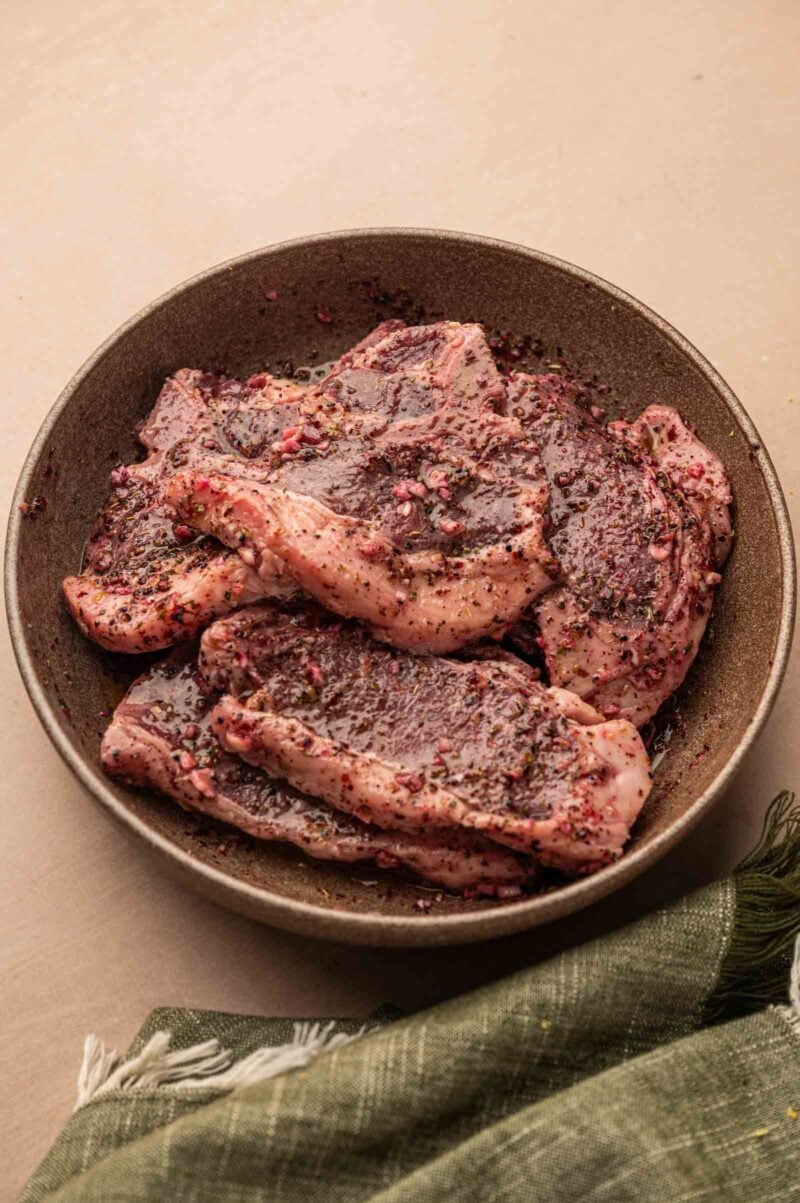 Raw lamb loin chops coated in a Greek lamb marinade are arranged in a brown bowl on a beige surface, with a green cloth partially visible nearby.