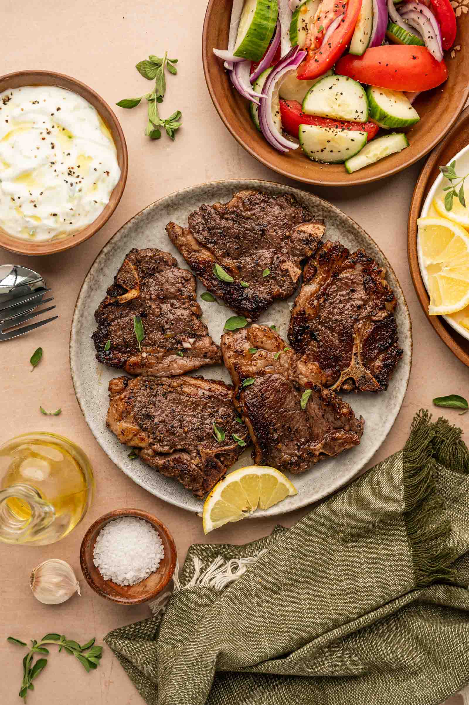A plate of seared lamb chops with a lemon wedge, surrounded by a Greek salad, lemon slices, tzatziki, olive oil, salt, and a green napkin.