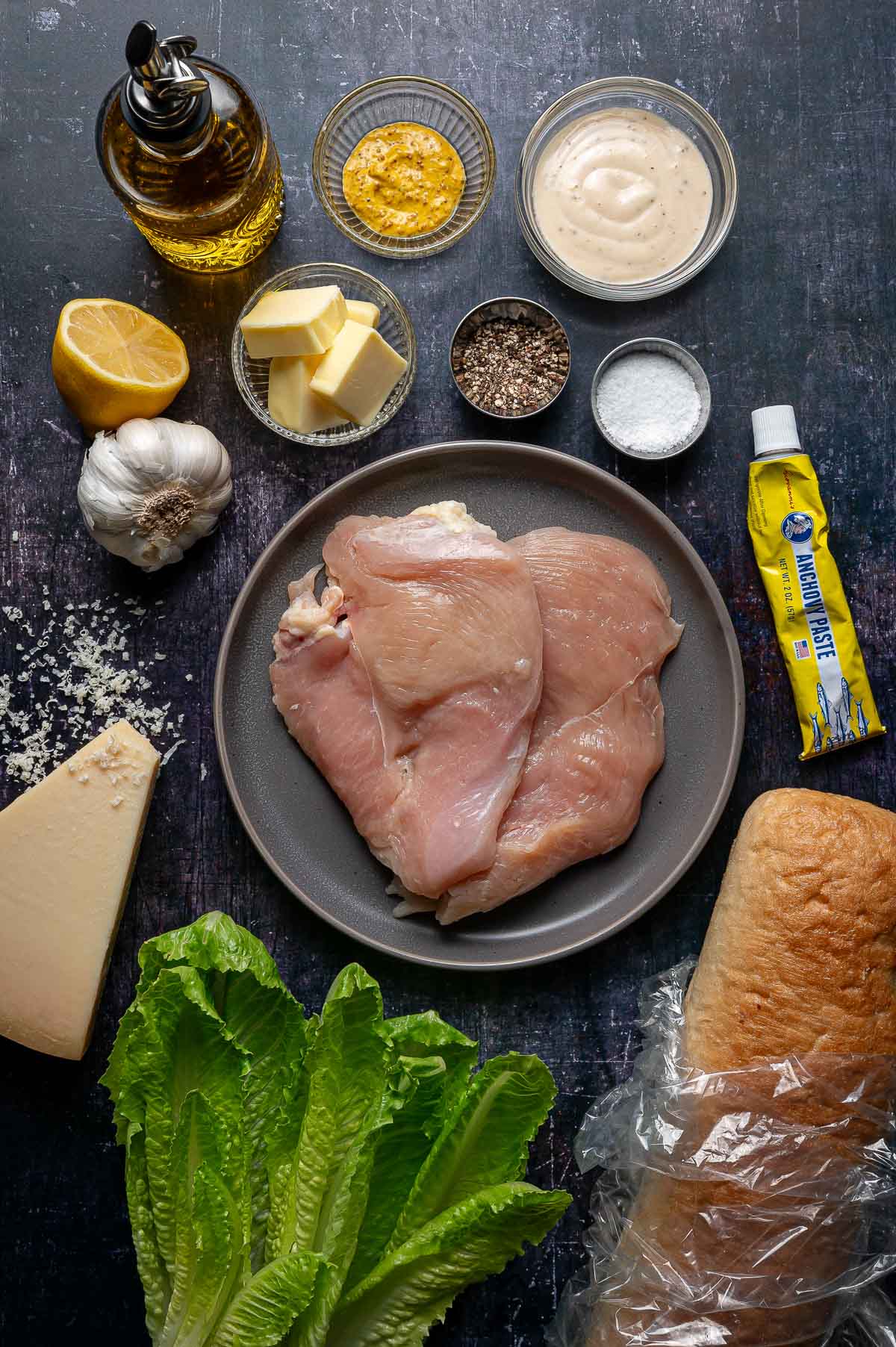 A flat lay of raw chicken breasts on a plate surrounded by romaine lettuce, bread, Parmesan cheese, garlic, lemon, butter, olive oil, mustard, black pepper, salt, mayonnaise, and anchovy paste.