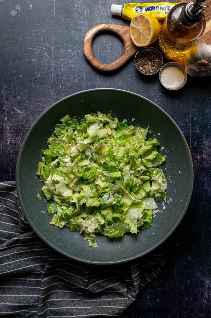 A bowl of chopped romaine lettuce, tossed in caesar dressing, with grated cheese, next to a board holding anchovy paste, lemon, salt, pepper, olive oil, and garlic.