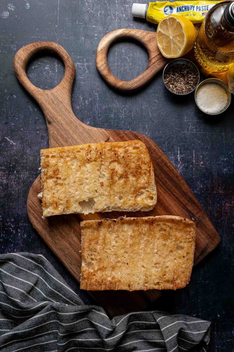 Two halves of toasted bread sit on a wooden cutting board, surrounded by a lemon, anchovy paste, oil, pepper, grated cheese, and a striped cloth.