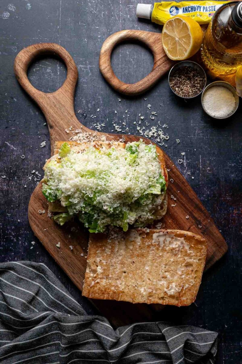 Open Caesar chicken sandwich on a wooden cutting board topped with lettuce and grated cheese, with seasonings, anchovy paste, lemon, and olive oil nearby. A striped cloth is on the lower edge.
