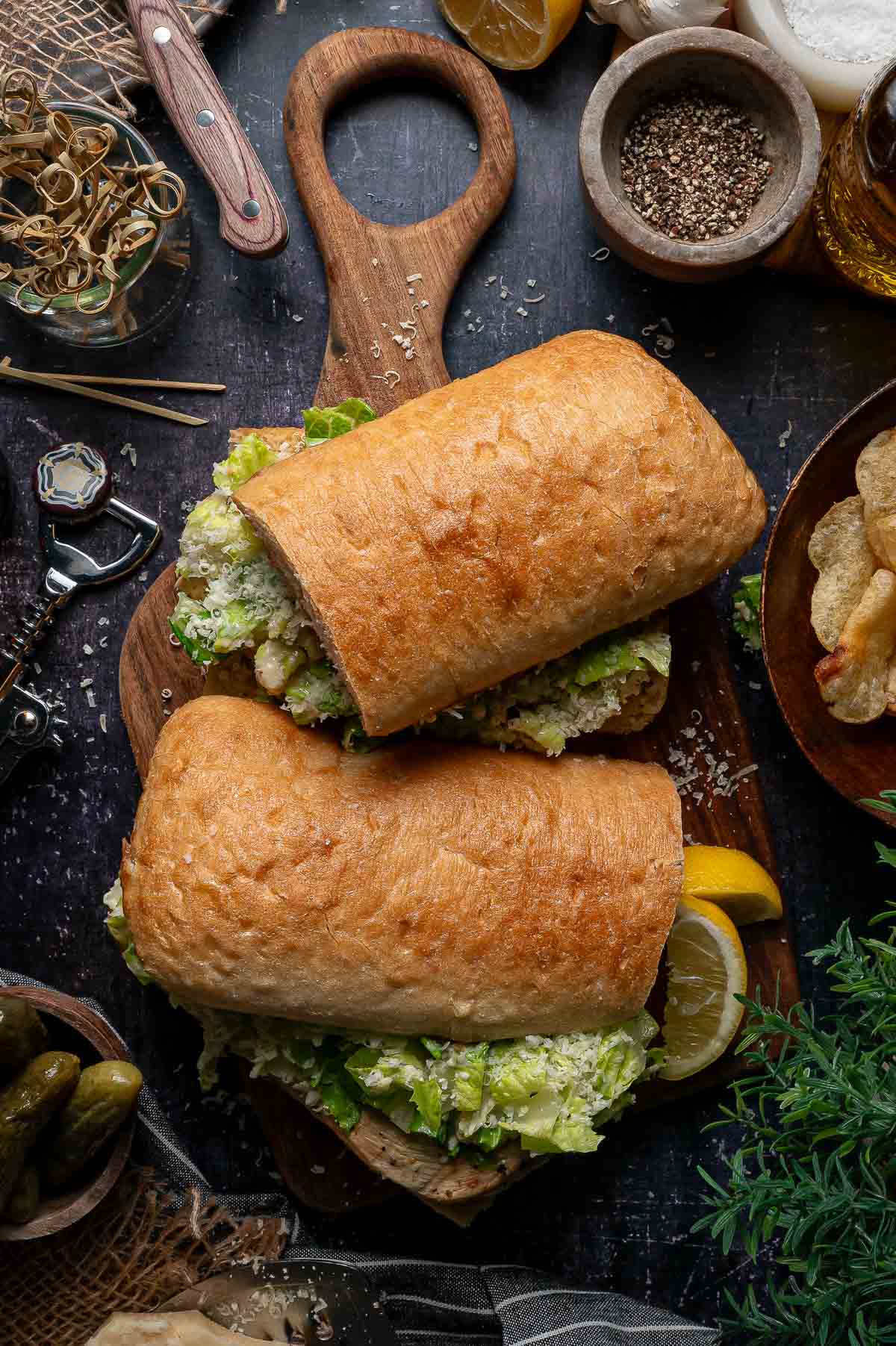 Two ciabatta rolls filled with grilled chicken caesar sit on a wooden board, surrounded by lemon wedges, pickles, potato chips, spices, and kitchen utensils.