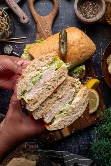 A hand holds a grilled chicken caesar sandwich sliced down the middle to show the filling, garnished with a pickle, on a wooden board with spices and a lemon slice nearby.