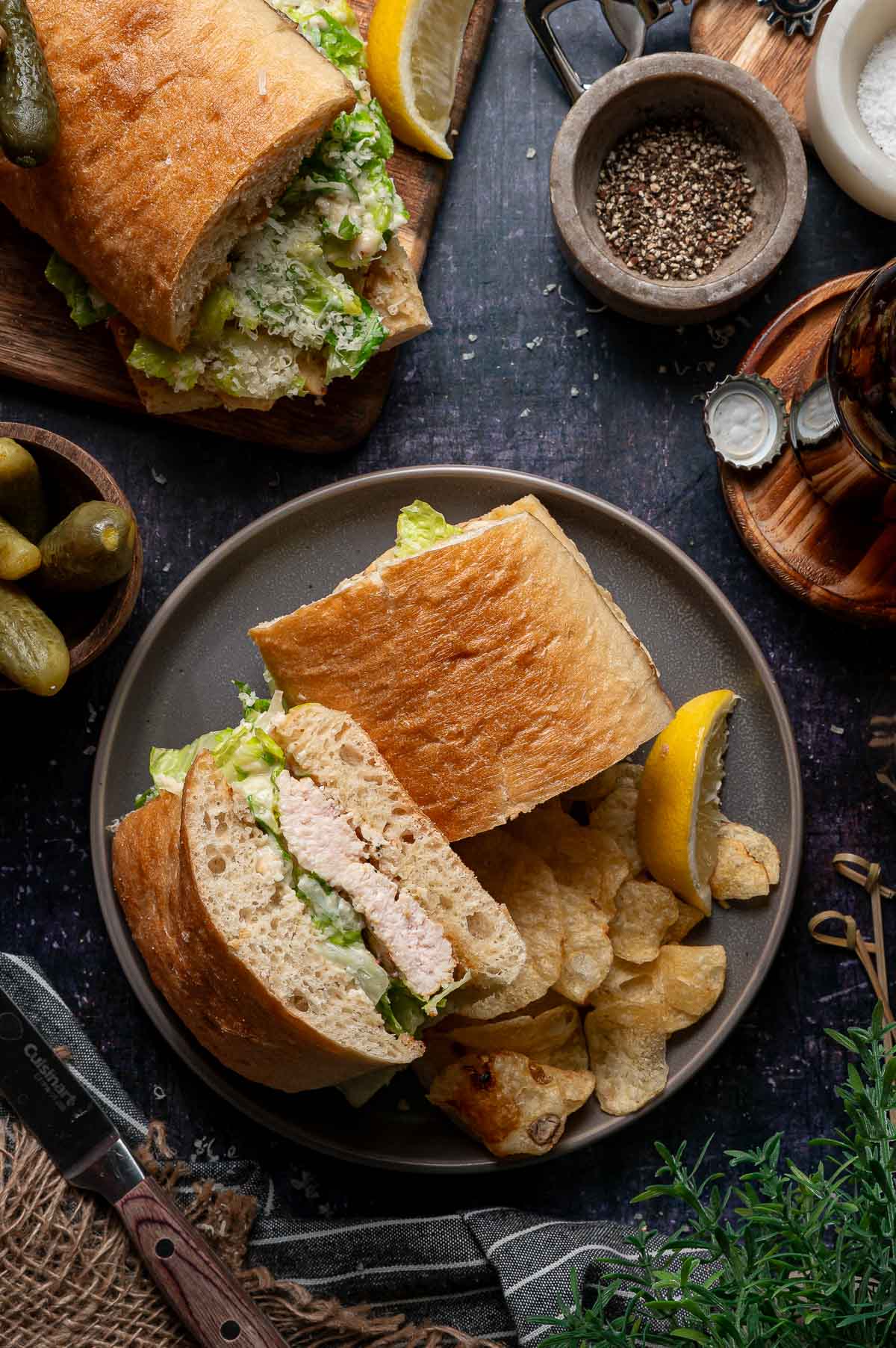 A plate with a grilled chicken caesar sandwich, a lemon wedge, potato chips, and pickles, with additional sandwich pieces, pepper, and condiments nearby on a dark surface.