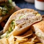 A grilled chicken caesar sandwich is cut in half on a plate, served with potato chips and a lemon wedge.