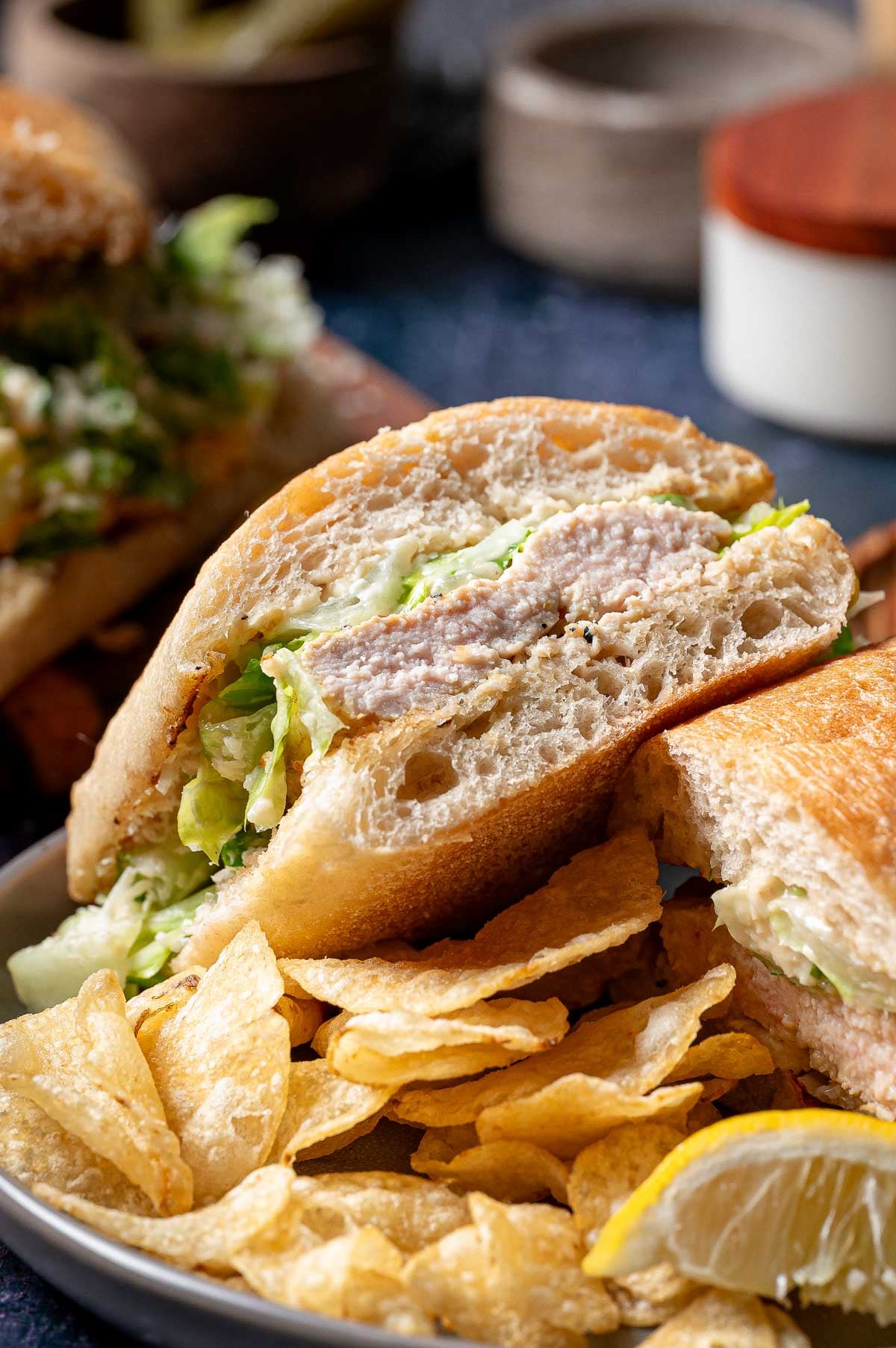 A grilled chicken caesar sandwich is cut in half on a plate, served with potato chips and a lemon wedge.
