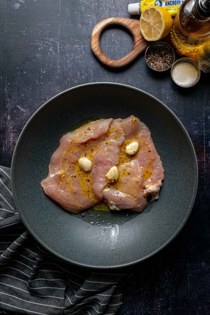 Raw chicken breasts on a dark plate, seasoned with olive oil, garlic cloves, and pepper. Lemon, anchovy paste, salt, pepper, and olive oil are visible nearby.