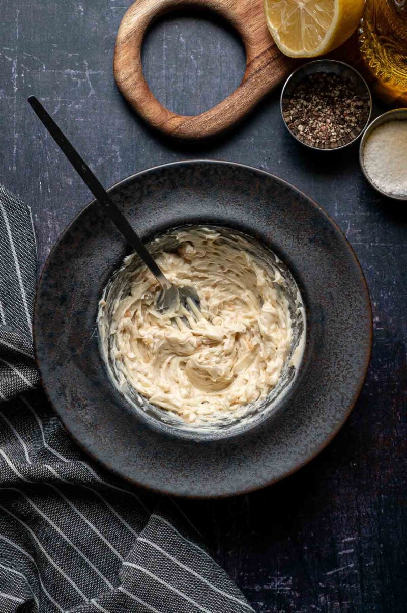 A bowl of seasoned anchovy butter is mixed with a fork, surrounded by a striped towel, lemon halves, black pepper, and grated cheese on a dark surface.