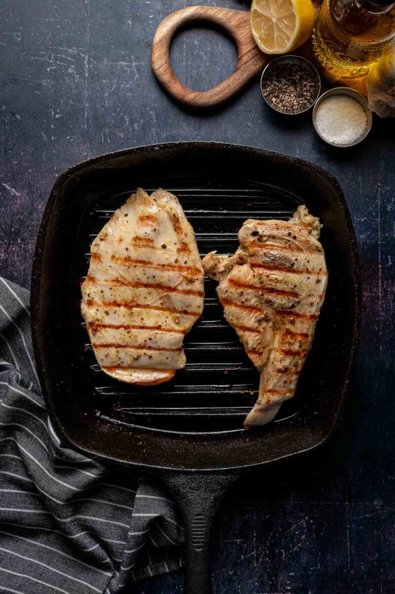 Two grilled chicken breasts in s grill pan , with seasoning ingredients and lemon above on a dark surface.