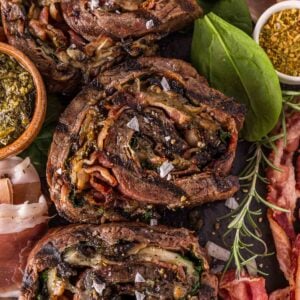 Three slices of rolled and stuffed flank steak with visible herbs, spinach, and bacon, garnished with salt and surrounded by fresh spinach and seasoning.