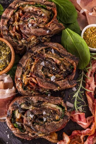 Three slices of rolled and stuffed flank steak with visible herbs, spinach, and bacon, garnished with salt and surrounded by fresh spinach and seasoning.