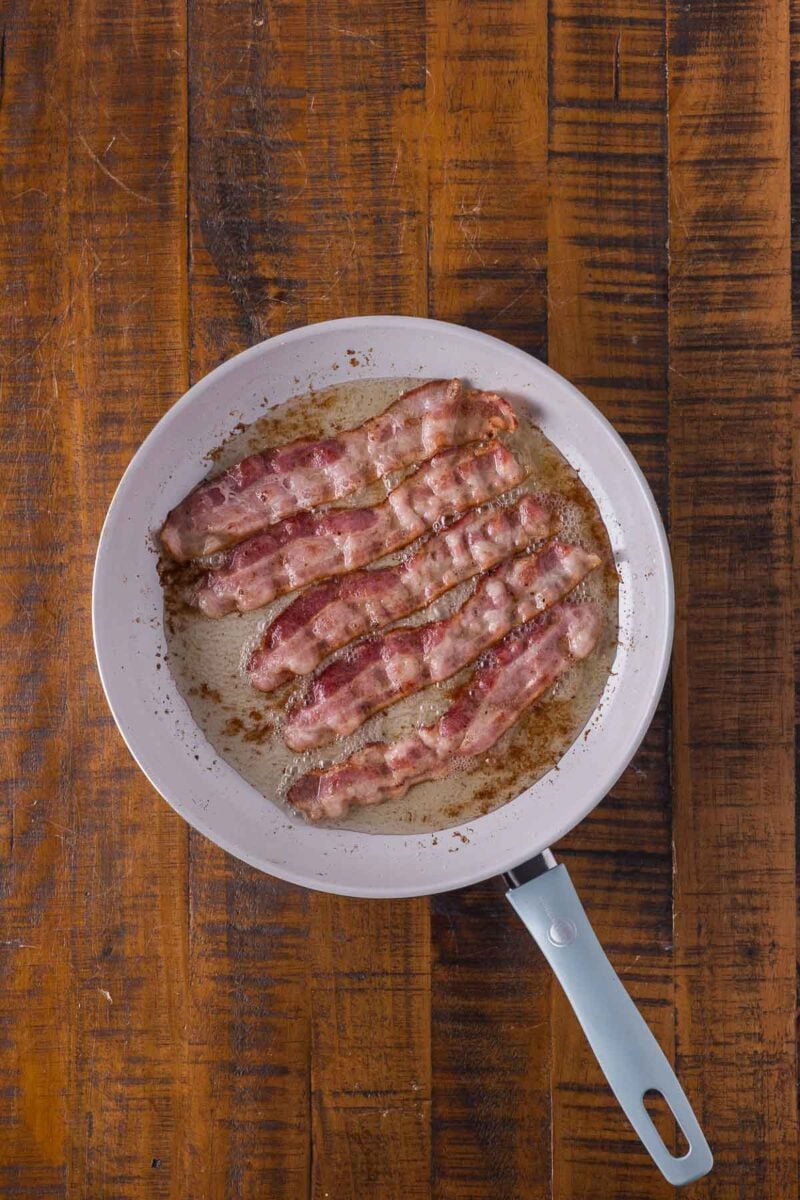 Six strips of bacon cooking in a white frying pan on a wooden surface.