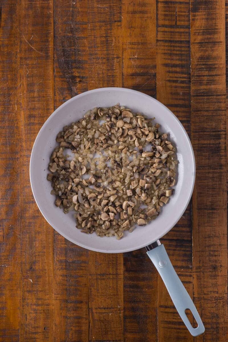 A white frying pan with chopped mushrooms and onions cooking in oil on a wooden surface.