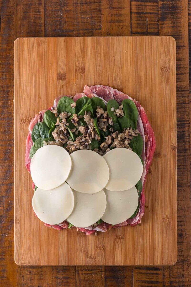 A wooden cutting board holds a butterflied flank steak layered with spinach, sauteed mushroom filling, and six slices of cheese.
